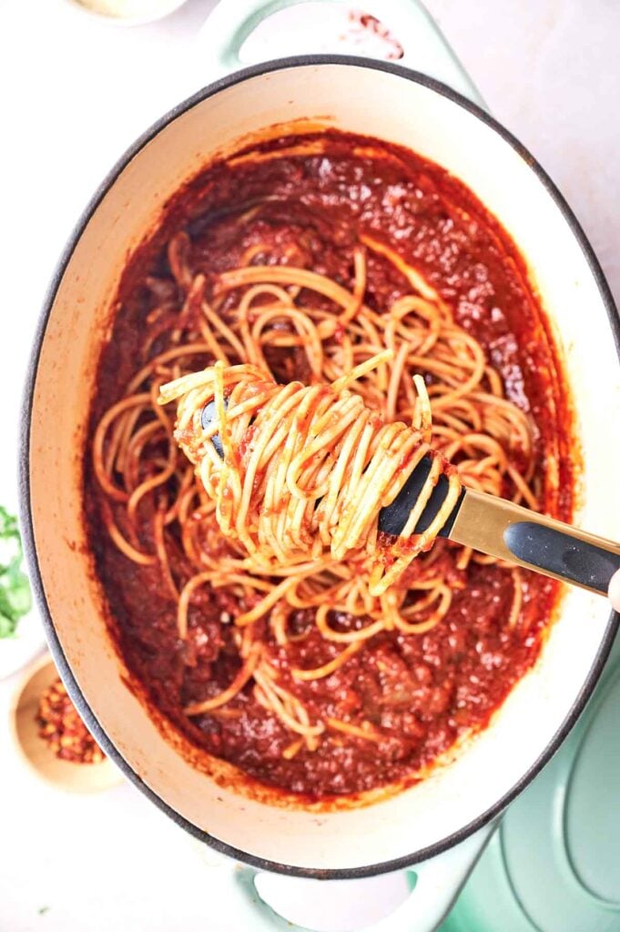 A pot of cooked Spaghetti Marinara mixed with rich tomato sauce, with a serving of pasta being lifted out using tongs.