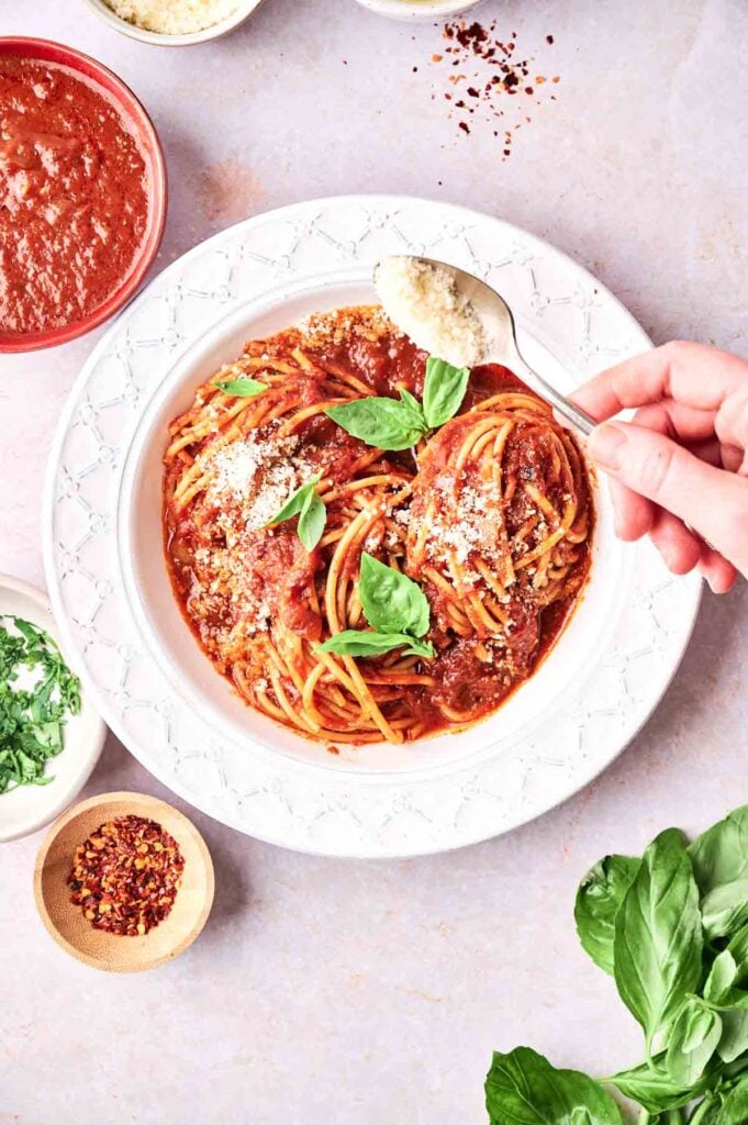 A hand sprinkles grated cheese over a plate of spaghetti marinara with tomato sauce and basil. Surrounding dishes hold extra sauce, red pepper flakes, grated cheese, and chopped herbs.