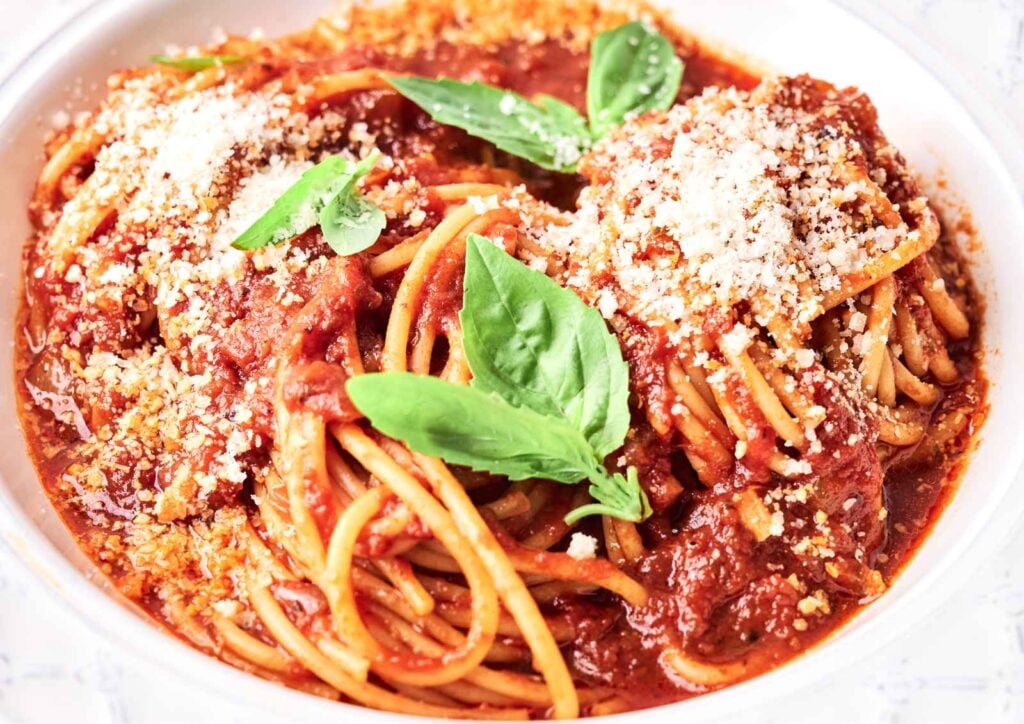 A plate of spaghetti marinara with tomato sauce, meatballs, grated cheese, and fresh basil leaves on top.