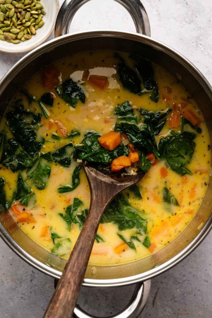 A pot of creamy Sweet Potato Soup with spinach and sweet potatoes, stirred with a wooden spoon, sits on a light surface next to a small bowl of pumpkin seeds.