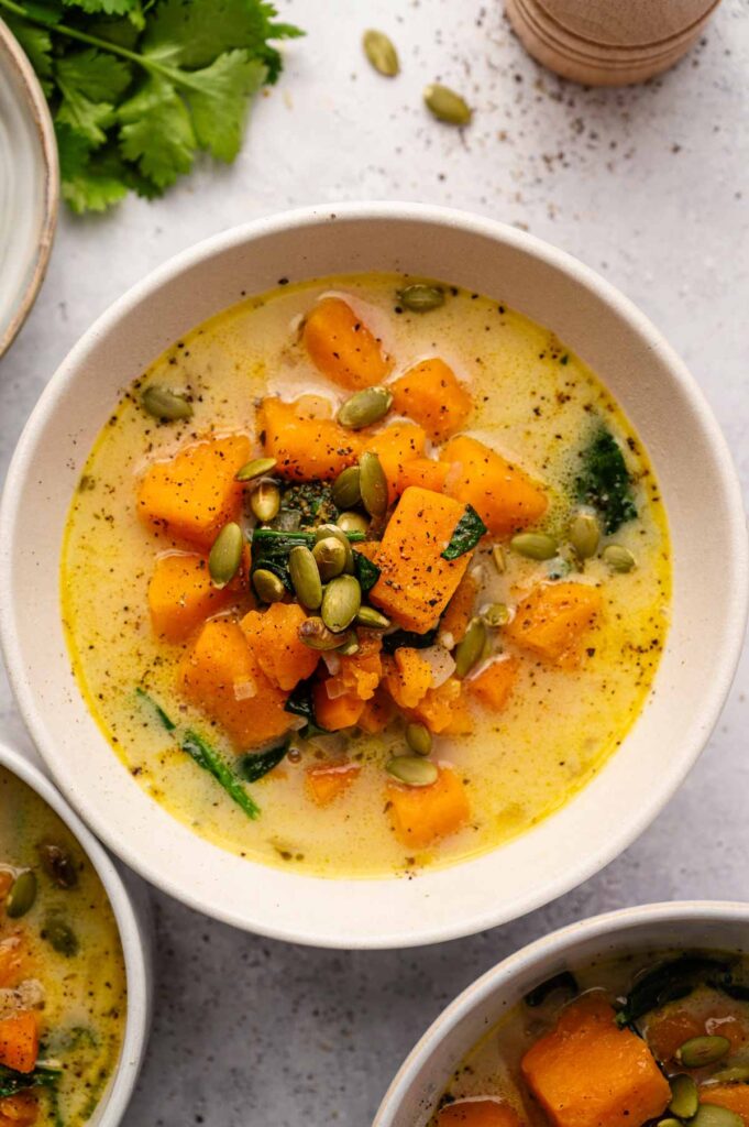 Bowl of creamy sweet potato soup with cubed orange squash, green leafy vegetables, and pumpkin seeds, garnished with black pepper.