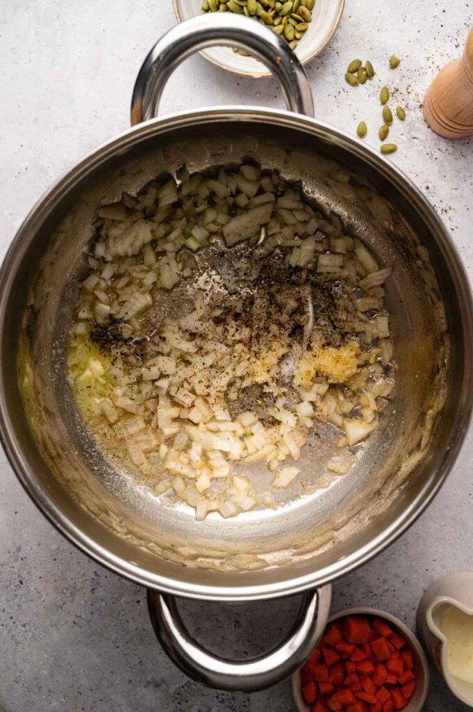 Stainless steel pot with chopped onions, garlic, salt, and pepper being sautéed for sweet potato soup; bowls of pumpkin seeds, carrots, and a pepper mill nearby.