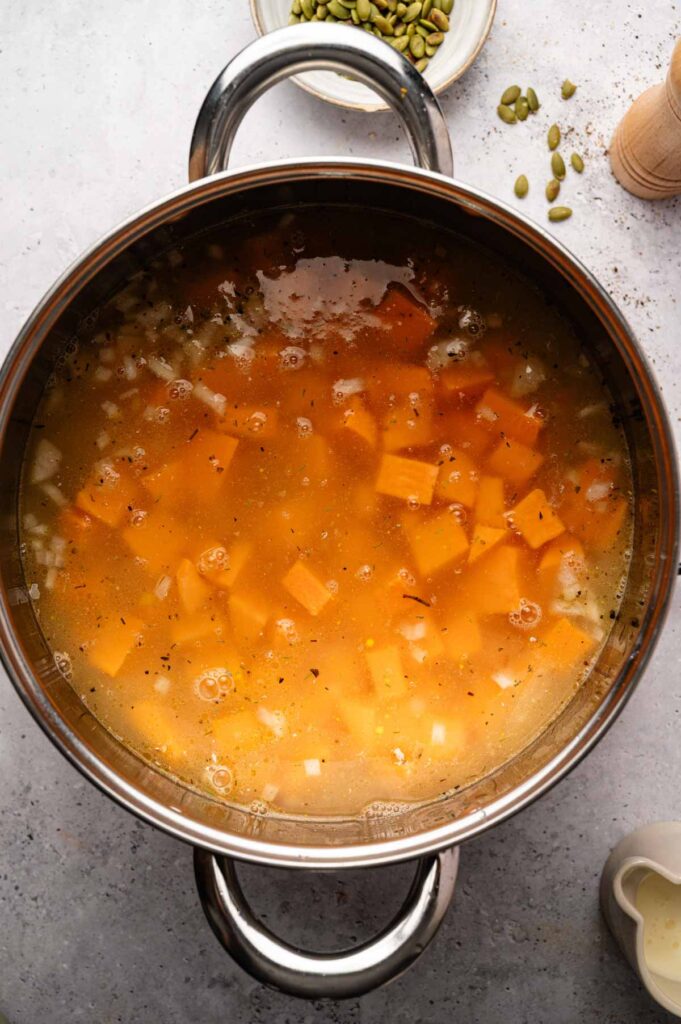 A stainless steel pot on a gray surface contains diced butternut squash, onions, and broth—the perfect base for a delicious sweet potato soup. Nearby are pumpkin seeds, a pepper grinder, and a small pitcher.