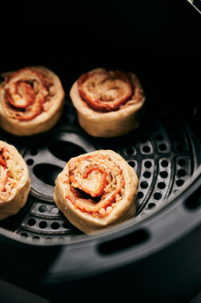 Four uncooked Air Fryer Pizza Rolls with visible cheese and sauce sit in an air fryer basket, ready to be cooked.