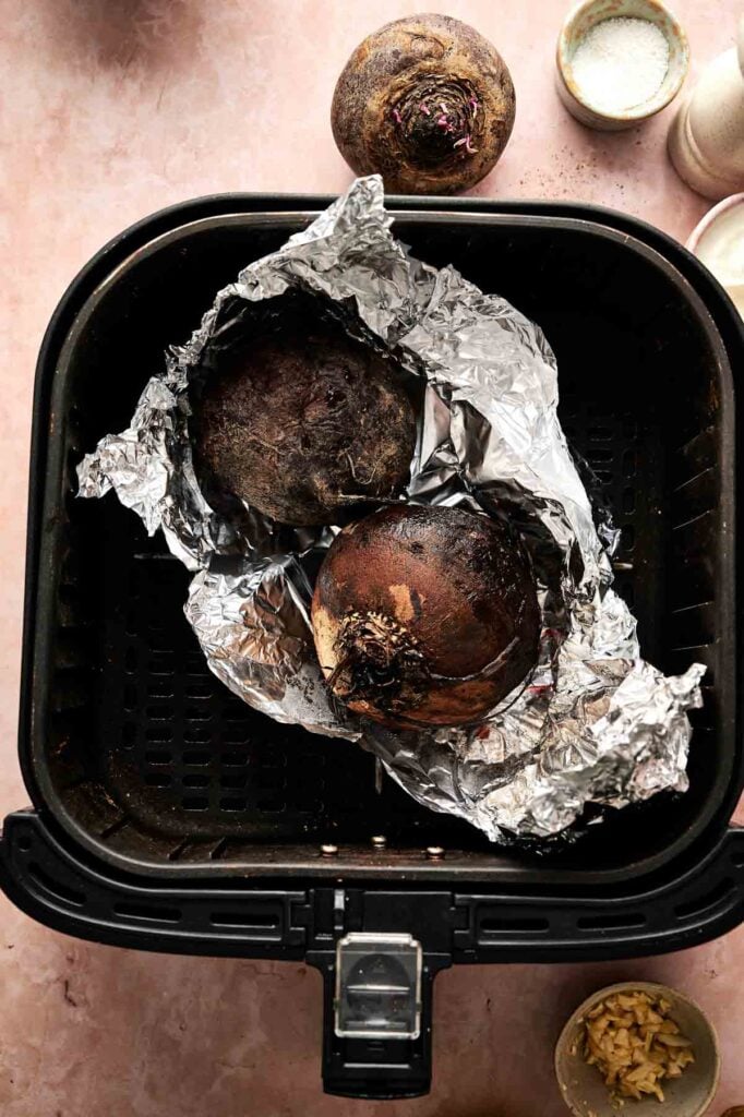Two whole beets wrapped in foil sit in an air fryer basket, with one unwrapped beet, a bowl of salt, and minced garlic&mdash;perfect prep for making delicious Air Fryer Beet Soup.