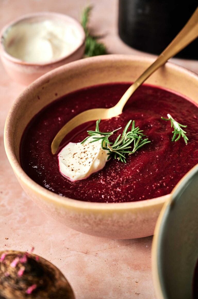 A bowl of Air Fryer Beet Soup garnished with a dollop of cream, a sprig of rosemary, and a spoon, with another small bowl in the background.