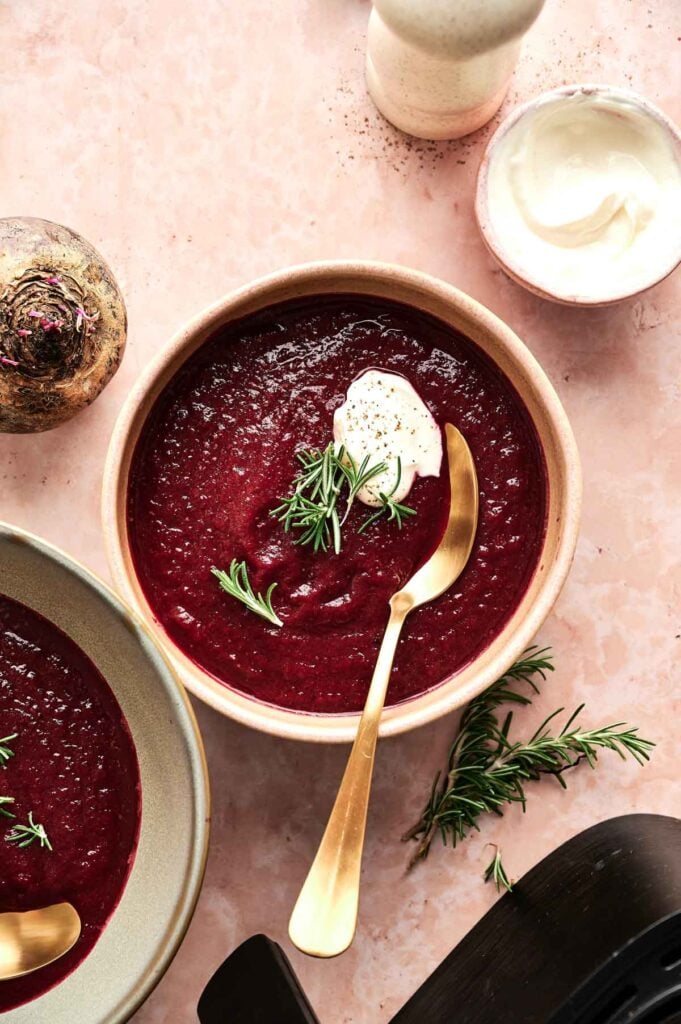 A bowl of Air Fryer Beet Soup topped with cream and a rosemary sprig, with a gold spoon resting inside. Nearby are a beet, a bowl of cream, and extra rosemary on a pink surface.