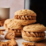 Four Air Fryer Biscoff Sandwich Cookies with a creamy filling are stacked on parchment paper, surrounded by broken cookie pieces and cups in the background.