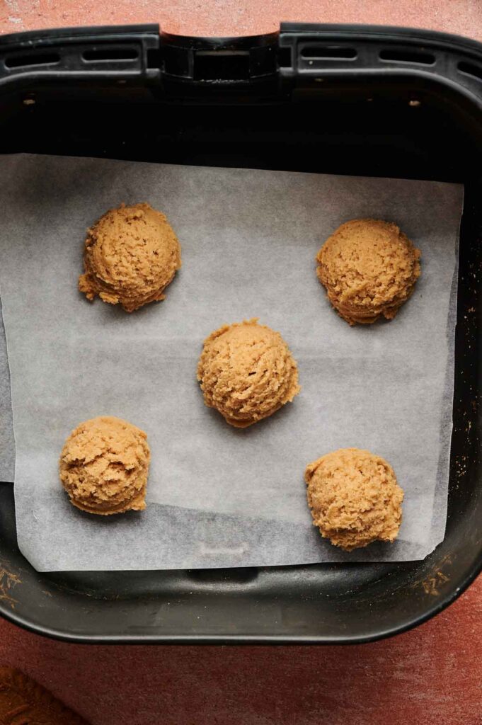 Five scoops of cookie dough for Air Fryer Biscoff Sandwich Cookies are placed on a sheet of parchment paper inside an air fryer basket, ready for baking.