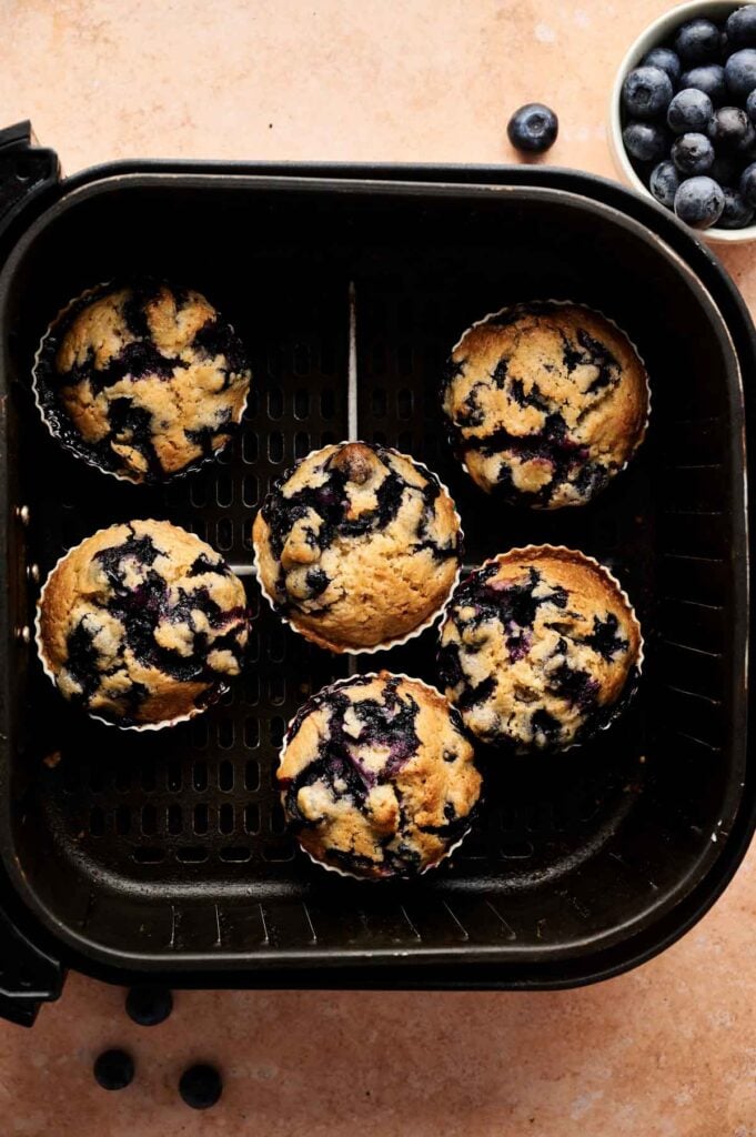 Six Air Fryer Blueberry Muffins in paper liners are arranged in an air fryer basket, with a bowl of fresh blueberries and a few loose berries nearby on a light surface.