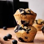 Two Air Fryer Blueberry Muffins are stacked on a table, surrounded by fresh blueberries, with another muffin in the background.