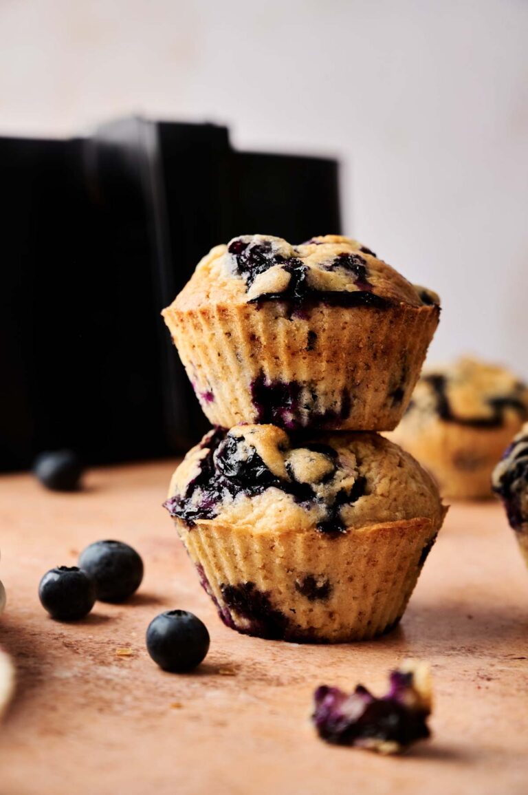 Two Air Fryer Blueberry Muffins are stacked on a table, surrounded by fresh blueberries, with another muffin in the background.