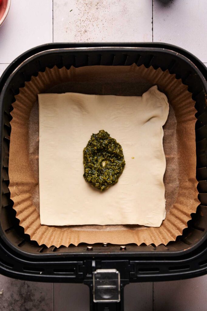 A square of puff pastry with a dollop of green pesto sits on parchment paper inside an air fryer basket, coming together to create a delicious Air Fryer Pesto Pizza in minutes.