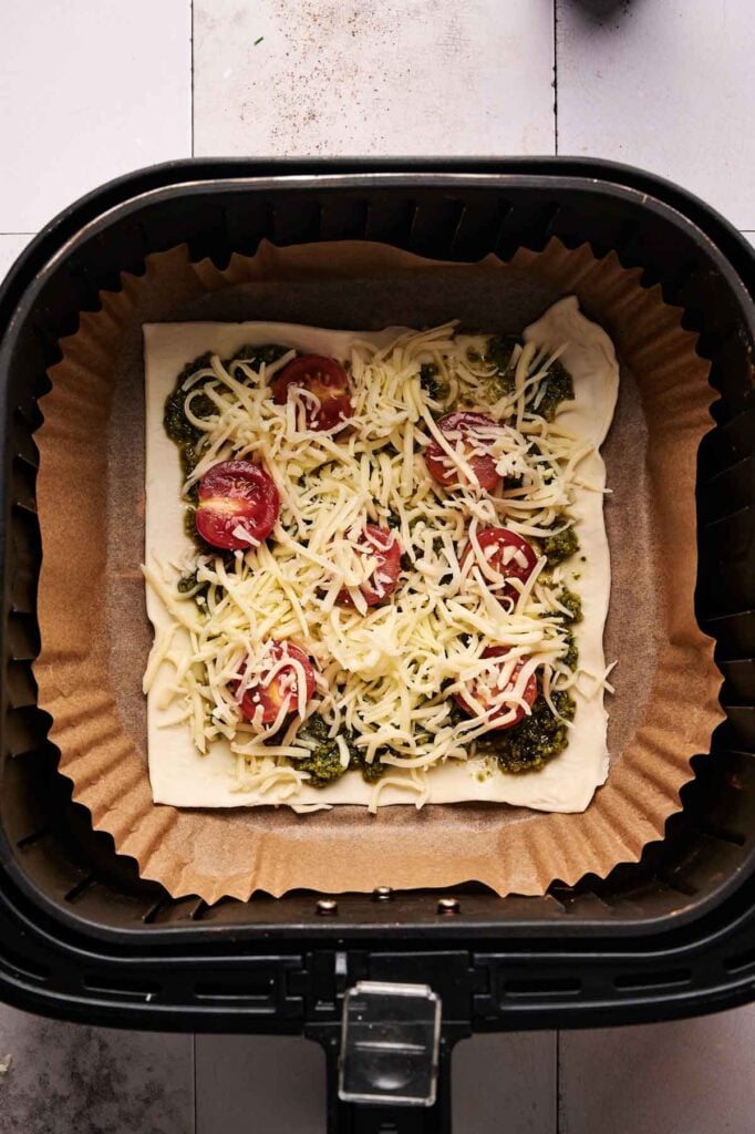 Air Fryer Pesto Pizza features unbaked puff pastry topped with pesto, halved cherry tomatoes, and shredded cheese, all arranged on parchment paper inside an air fryer basket.