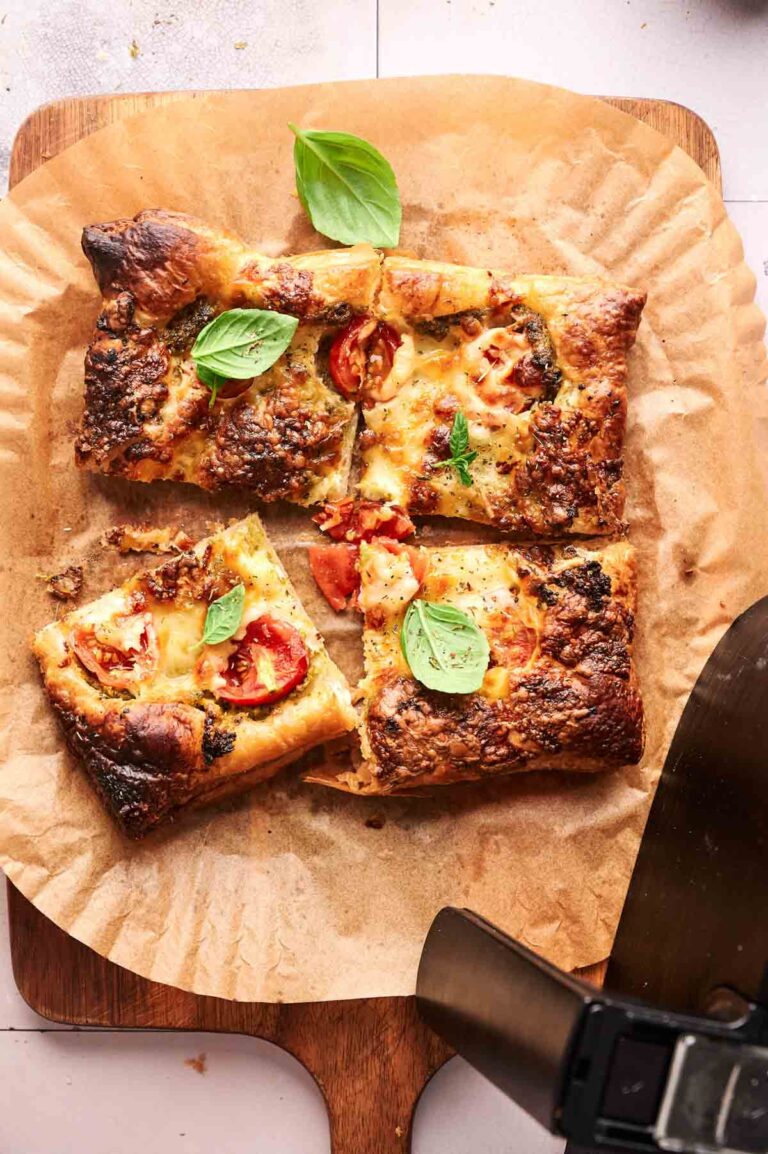 A rectangular Air Fryer Pesto Pizza with melted cheese, tomatoes, and basil leaves, cut into four pieces on parchment paper atop a wooden board.