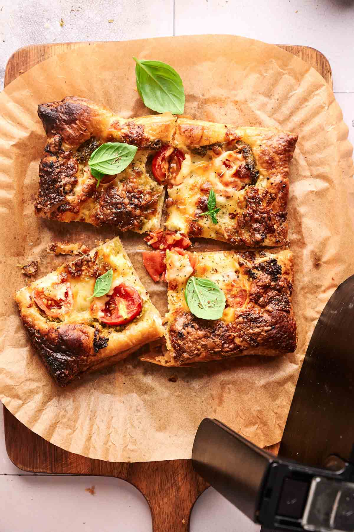 A rectangular Air Fryer Pesto Pizza with melted cheese, tomatoes, and basil leaves, cut into four pieces on parchment paper atop a wooden board.