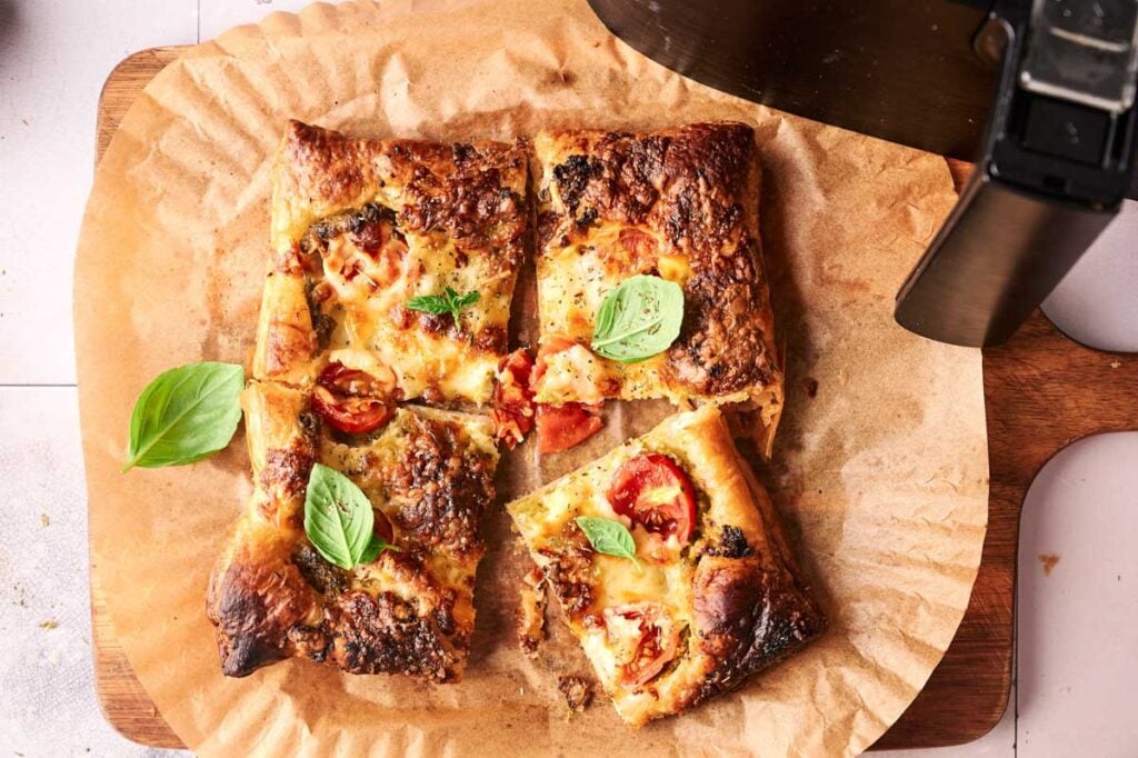 Rectangular Air Fryer Pesto Pizza with melted cheese, tomatoes, and basil leaves, cut into four pieces on parchment paper atop a wooden board.