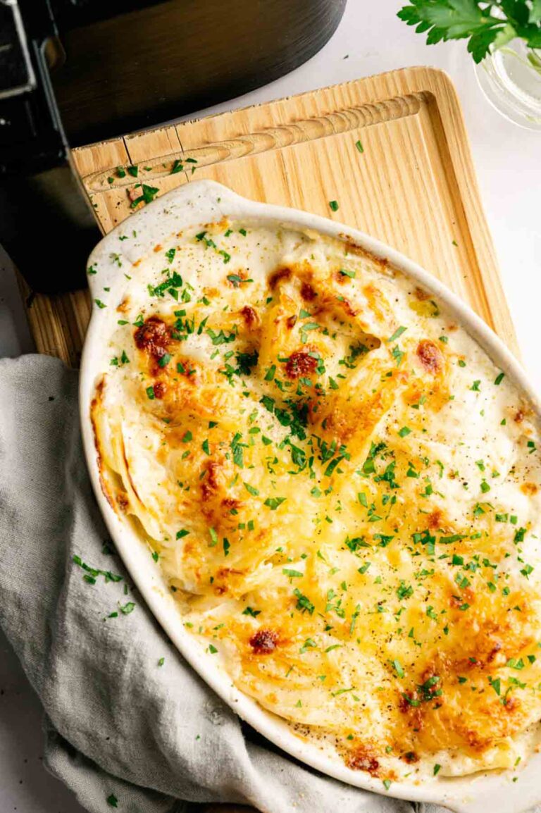 A baked dish of cheesy Air Fryer Potatoes Au Gratin garnished with chopped parsley, served in an oval white casserole dish on a wooden board.