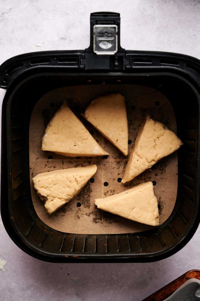 Five unbaked Air Fryer Scones wedges are arranged on parchment paper inside an air fryer basket, ready to be baked.