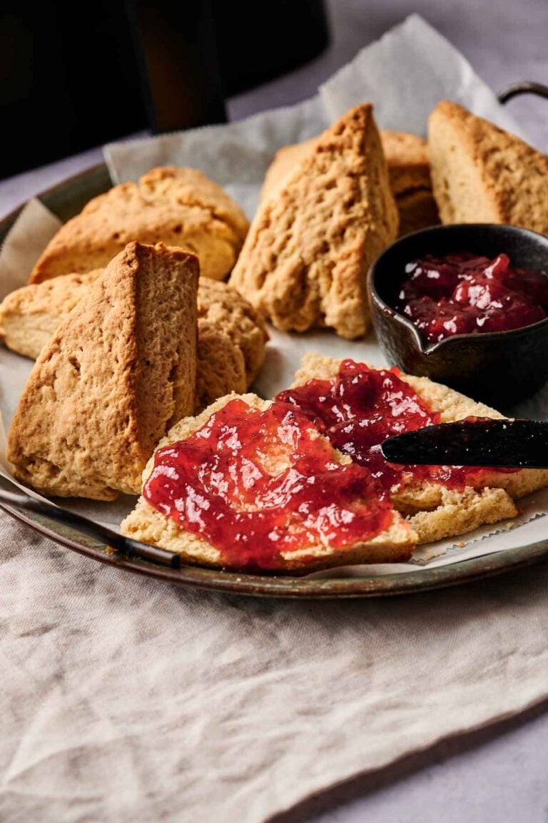 Plate of Air Fryer Scones, one halved and spread with red jam, with a small bowl of jam and a knife on a cloth-covered surface.