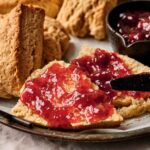 A close-up of an Air Fryer Scone split in half and spread with red jam, with more air fryer scones and a dish of jam in the background.