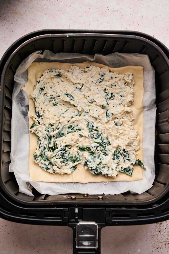 Rectangular pastry dough with a spinach and cheese mixture spread on top, placed on parchment paper inside an air fryer basket, creates a quick and delicious Air Fryer Spinach Pizza.