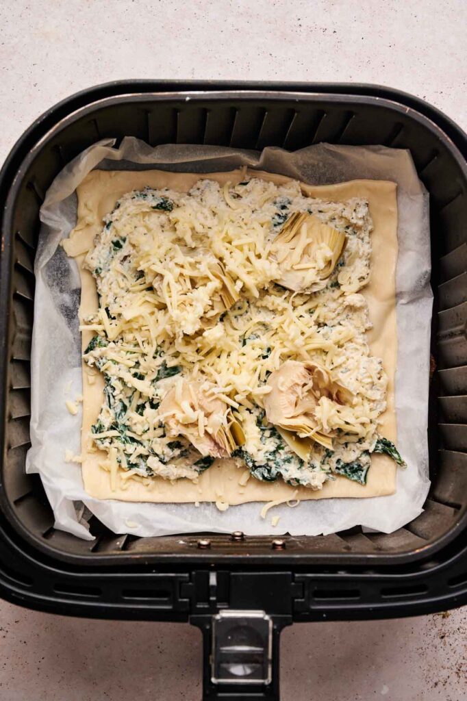 Unbaked pastry topped with spinach, cheese, artichoke, and creamy filling sits on parchment paper inside an air fryer basket, ready to be transformed into a delicious Air Fryer Spinach Pizza.