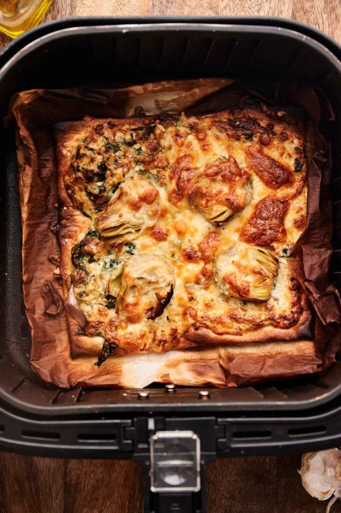 A baked savory tart, reminiscent of an Air Fryer Spinach Pizza, features melted cheese, artichoke hearts, and greens atop parchment paper inside an air fryer basket.