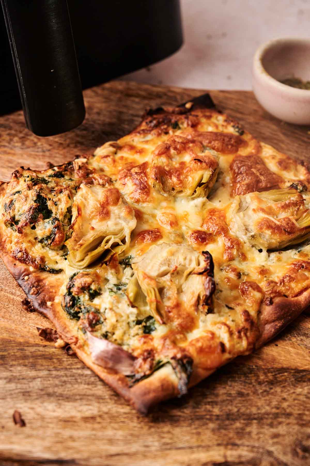 An Air Fryer Spinach Pizza features a baked flatbread topped with melted cheese, artichoke hearts, and spinach, displayed on a wooden cutting board.