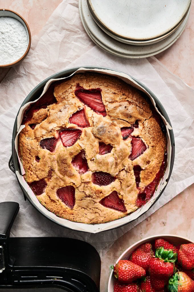 An Air Fryer Strawberry Cake in a round pan sits on parchment paper, with plates, a bowl of strawberries, and a bowl of powdered sugar nearby.