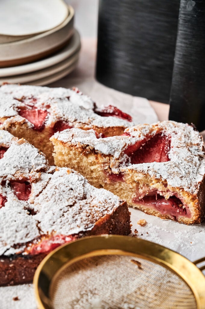 An Air Fryer Strawberry Cake, sliced and dusted with powdered sugar, is displayed on parchment paper, with plates stacked in the background and a mesh strainer in the foreground.