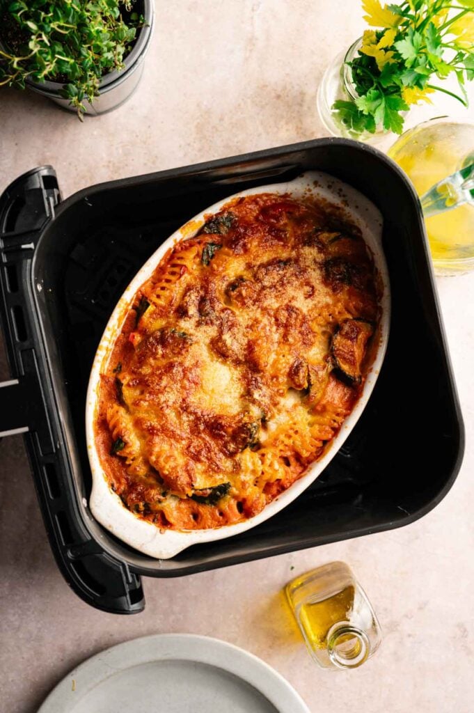 An Air Fryer Vegetable Pasta Bake sits in an oval dish, topped with melted cheese and surrounded by fresh herbs, a drink, and a small bottle of olive oil.