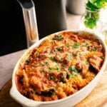 Air Fryer Vegetable Pasta Bake casserole with cheese and tomato sauce in a white oval dish, garnished with herbs, placed on a wooden board next to an air fryer and potted plants.