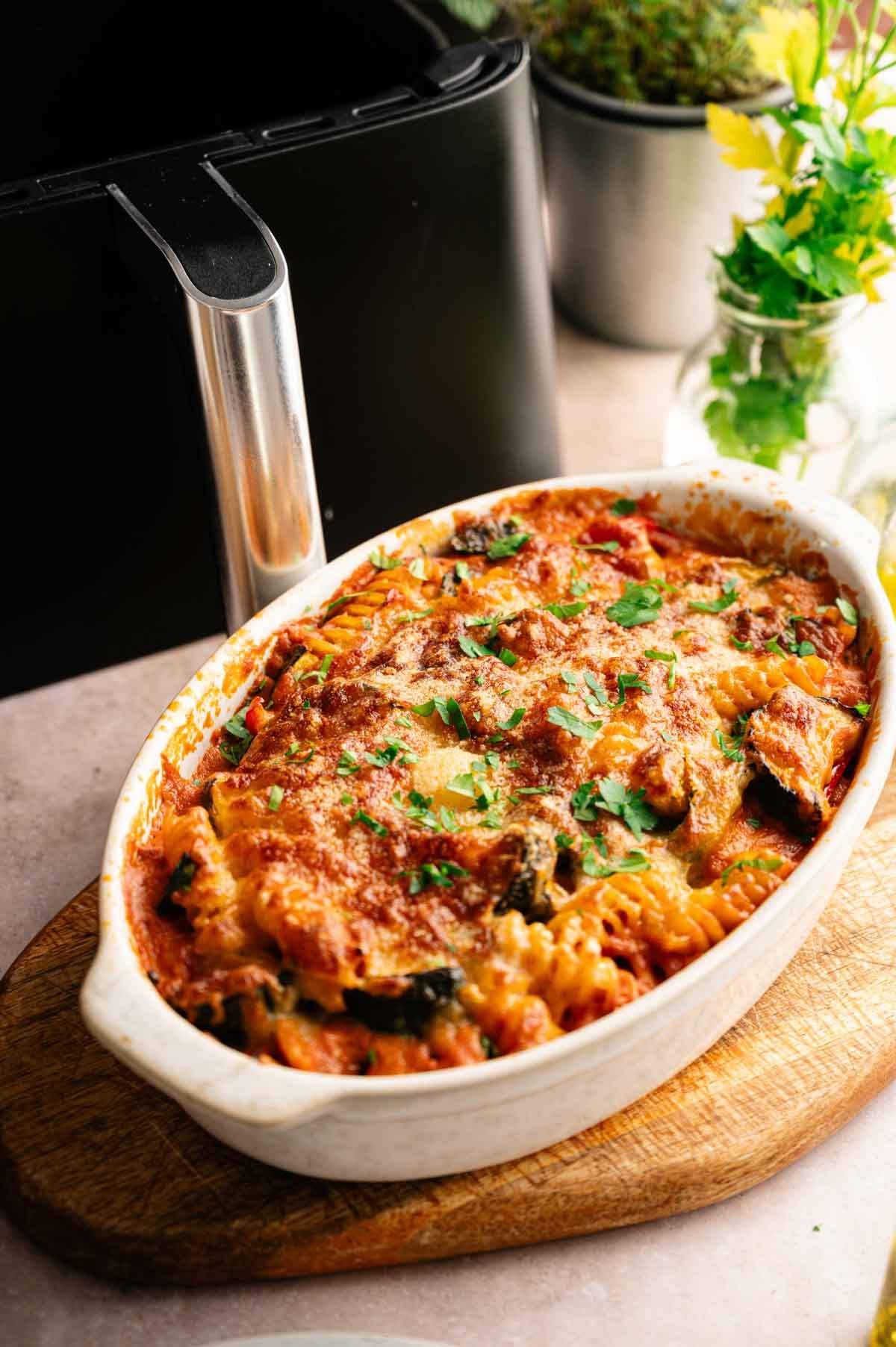 An Air Fryer Vegetable Pasta Bake casserole topped with melted cheese and herbs, served in a white oval dish on a wooden board, sits next to an air fryer and some potted plants.