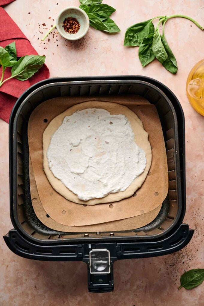 Uncooked Air Fryer White Pizza dough topped with creamy white sauce sits on parchment paper inside an air fryer basket, surrounded by fresh basil, a red napkin, and a small bowl of red pepper flakes.
