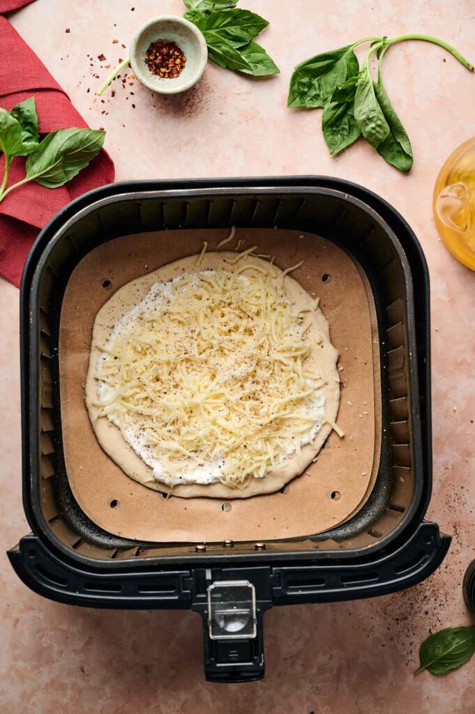 An uncooked Air Fryer White Pizza topped with cheese sits on parchment paper inside an air fryer basket, surrounded by fresh basil, a red cloth, and a bowl of chili flakes on a countertop.
