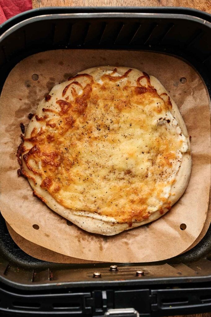An Air Fryer White Pizza, featuring a baked flatbread topped with melted cheese, sits on parchment paper inside an air fryer basket.