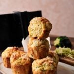 A stack of five savory Air Fryer Zucchini Muffins on a beige plate, with one muffin broken open to reveal its moist inside; grated zucchini and a box grater are in the background.