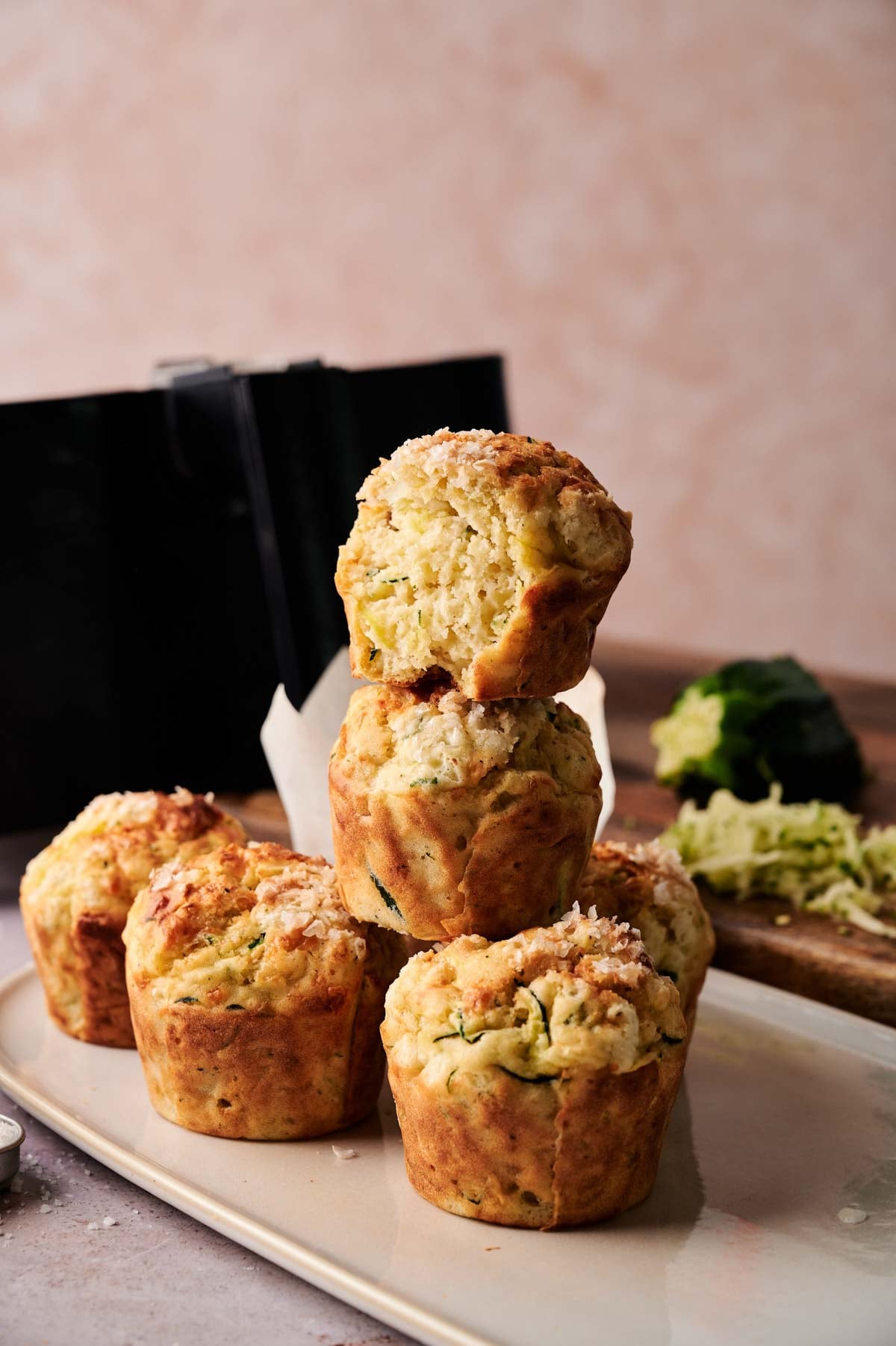 A stack of five savory Air Fryer Zucchini Muffins on a beige plate, with one muffin broken open to reveal its moist inside; grated zucchini and a box grater are in the background.