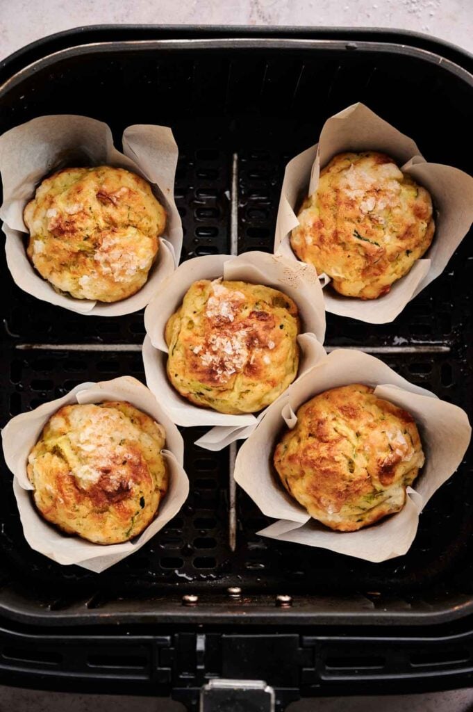 Five Air Fryer Zucchini Muffins in parchment paper cups sit inside an air fryer basket.