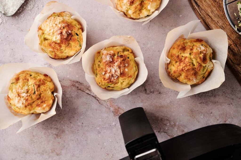 Five Air Fryer Zucchini Muffins in white parchment paper cups are arranged on a light marble surface, with part of an air fryer visible in the foreground.