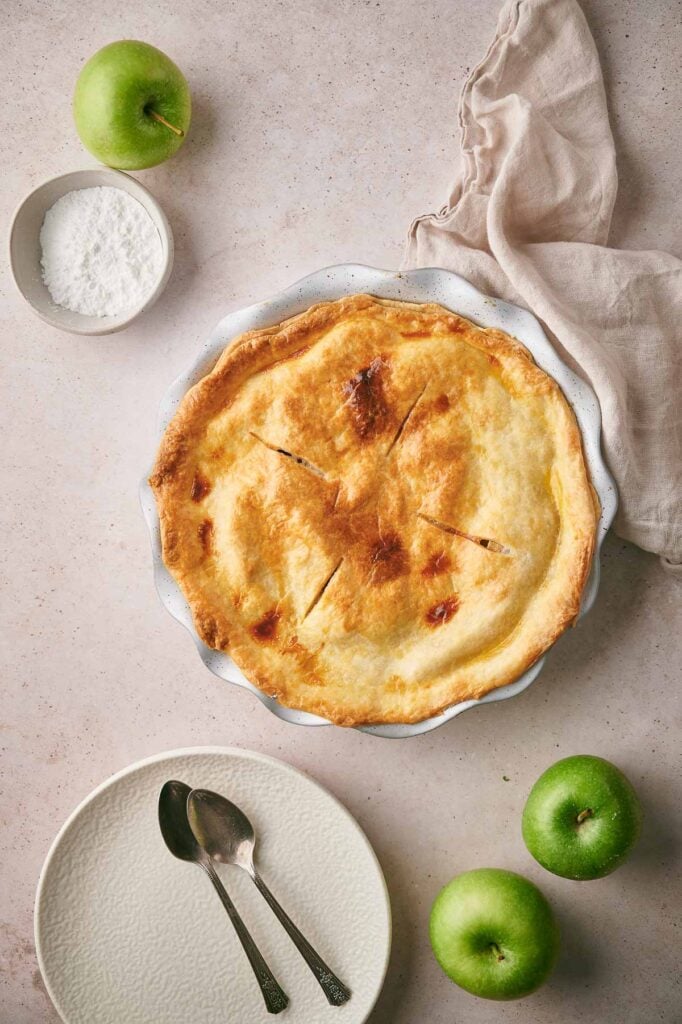 Overhead view of a freshly baked apple pie in a dish, surrounded by three green apples, a bowl of powdered sugar, a beige cloth, and an empty plate with two spoons.