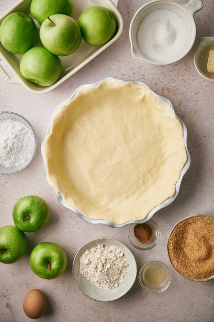Overhead view of an empty pie crust in a dish surrounded by green apples, sugar, flour, spices, an egg, powdered sugar, lemon juice, and a block of butter—everything you need to create the perfect apple pie.