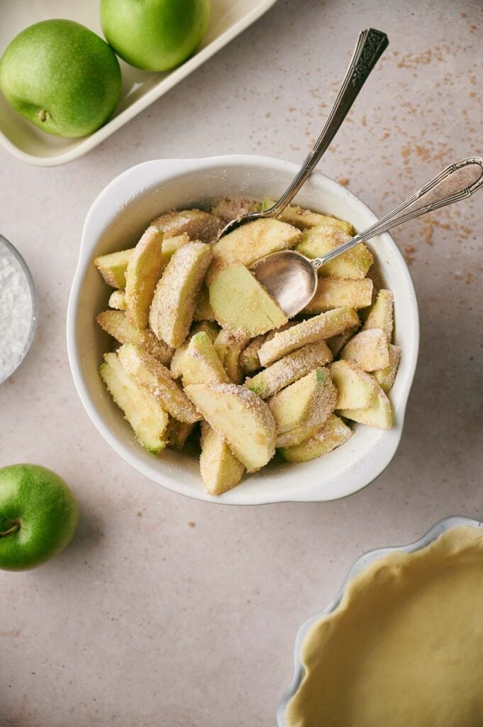 Sliced green apples coated with sugar and cinnamon in a bowl with two spoons, surrounded by whole apples, powdered sugar, and an empty pie crust—perfect ingredients for a homemade apple pie.