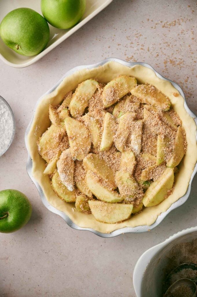 Unbaked apple pie with sliced green apples and cinnamon sugar in a pie crust, surrounded by whole green apples and classic apple pie baking ingredients on a countertop.