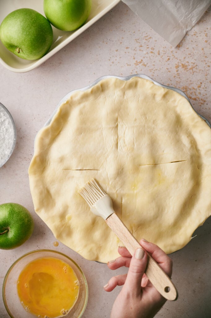 A hand uses a pastry brush to apply egg wash to an unbaked apple pie with slits in the crust; green apples and a bowl of beaten egg are nearby.