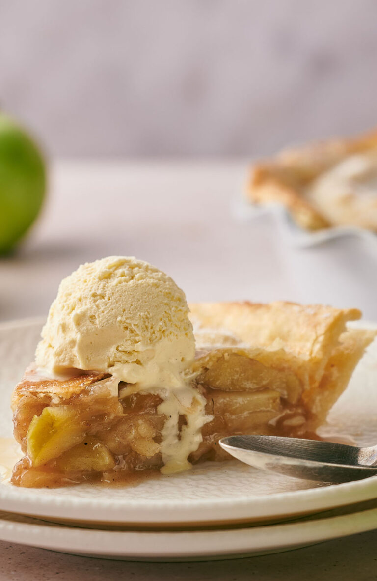 A slice of classic Apple Pie topped with vanilla ice cream sits on a plate with a fork, while a green apple and a pie dish add a cozy touch in the background.
