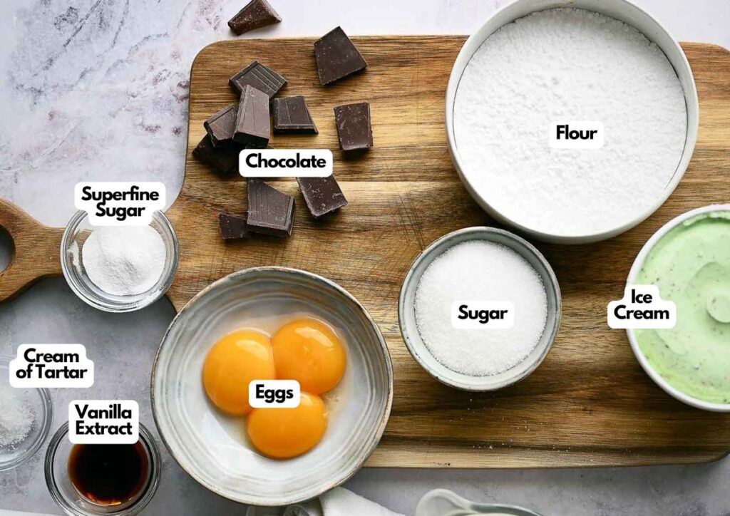 A wooden board holds all the labeled ingredients for Baked Alaska: bowls of flour, sugar, ice cream, chocolate pieces, egg yolks, cream of tartar, superfine sugar, and vanilla extract.