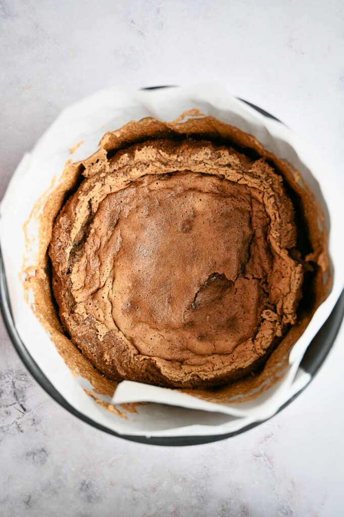 A baked chocolate cake with a cracked top sits in a round pan lined with parchment paper, reminiscent of a classic Baked Alaska, viewed from above.