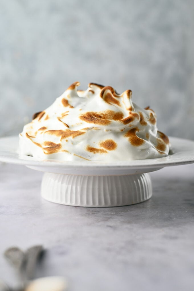 A classic Baked Alaska dessert with beautifully toasted meringue sits on a white cake stand against a light, textured background.
